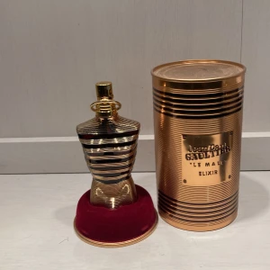 Jean Paul Gaultier Le Male Elixir - Exklusiv parfym i guldig, metallisk flaska formad som en manlig torso. Kommer med en lyxig röd sammetsbas och matchande guldfärgad burk med präglad logga. Doften är intensiv och modern, perfekt för dig som vill sticka ut.