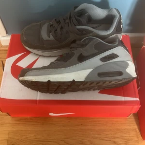 Nike Air Max 90 grå sneakers - Säljer ett par klassiska Nike Air Max 90 sneakers i olika grå nyanser med svarta detaljer och vit sula. Skorna har den ikoniska Air Max-dämpningen och är tillverkade i en mix av syntet och skinn. Perfekta för dig som gillar streetwear och sportig stil.