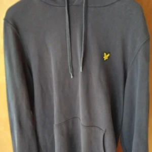Svart hoodie från Lyle & Scott - Svart hoodie från Lyle & Scott med klassisk gul logga på bröstet. Tröjan har huva med snörning och en stor magficka framtill. Mjuk bomullskvalitet och långärmad modell, perfekt för chill dagar. Snygg och enkel design som funkar till det mesta. (Liten i storleken passar M bättre)