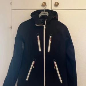Svart skidjacka från Tenson med huva - Svart skidjacka från Tenson med vit dragkedja och fyra snedställda fickor framtill. Jackan har huva, diskret logga på bröstet och är tillverkad i ett slätt syntetmaterial.