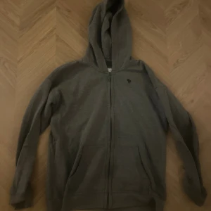 Grå hoodie med dragkedja från Abercrombie - Mysig grå hoodie från Abercrombie & Fitch med dragkedja framtill och klassisk huva. Tröjan har två fickor och en liten svart älg-logga broderad på bröstet. Väldigt bra kvalitet och sparsamt använd! Nypris är runt‼️ 1000kr‼️