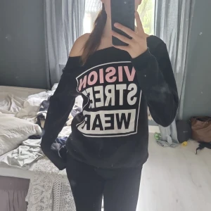 Svart offshoulder tröja från Elfin - Svart långärmad tröja från Elfin med offshoulder-modell och tryck i vitt och rosa framtill där det står 'VISION STREET WEAR'. Mjuk och skön, perfekt för en avslappnad streetstyle-look.