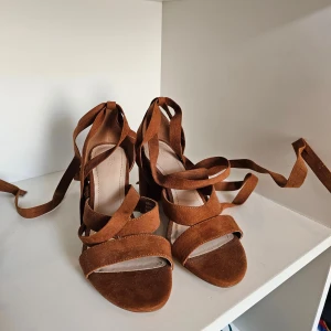 Bruna mockasandaler med klack och knyt från H&M - Snygga sandaler i brun mocka med bred klack och långa remmar att knyta runt vristen. Skorna har öppen tå och platt sula framtill, perfekt för dig som gillar en trendig och chic look. Passar till både jeans och klänning.