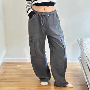 Grå cargopants från Bershka med snörning - Ett par grå cargopants från Bershka i storlek M. Byxorna har bred passform, snörning i midjan så storleken runt midjan går att anpassa. Slitna detaljer längst ner men även blivit slitna på baksidan längst ner. Fortfarande väldigt coola byxor!