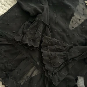 Svart broderad blus från H&M - Supersöt svart blus från H&M med broderade små blommor och volangdetaljer vid ärmslut och halsringning. Blusen har v-ringning och vida, långa ärmar med romantisk känsla. Perfekt för dig som gillar unika detaljer och flowy stil.