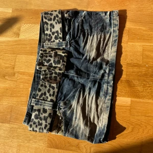 Jeanskjol med leopardbälte - Cool kort jeanskjol i blått med slitna detaljer och stenar framtill. Kjolen har ett brett bälte med leopardmönster och stor spänne som verkligen sticker ut. Fickor både fram och bak. Perfekt för dig som vill ha en edgy och unik stil.