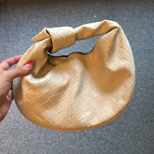 Beige axelväska med knut och rutmönster - Säljer en beige axelväska i syntetmaterial med rutmönstrad yta. Väskan har en rund form, en cool knutdetalj på handtaget och stängs med dragkedja. Perfekt storlek för det viktigaste och ger en trendig vibe till din outfit.