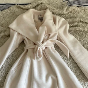 Beige ASOS kappa M - Snygg beige kappa från ASOS med bred krage och knytbälte i midjan. Jackan har långa ärmar. Storlek 40 står för M finns ett litet snit hål innanför jackan men påverkar inte hur den sitter och syns inte alls 