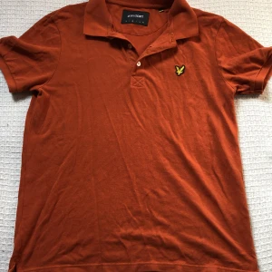 Orange Lyle & Scott pikétröja - Snygg orange pikétröja från Lyle & Scott i 100% bomull. Klassisk krage med två knappar och den ikoniska gula fågeln broderad på bröstet. Perfekt för dig som gillar stilrena och sportiga plagg med en cool touch.