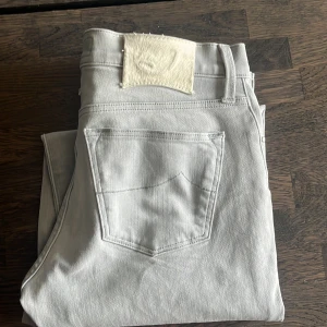 Ljusgrå jeans från Jacob Cohën, stl 28 - Snygga ljusgrå jeans från Jacob Cohën med klassisk femficksdesign och exklusiv etikett i pälsimitation bak. Byxorna är raka i modellen och tillverkade i mjukt bomullstyg med en lyxig känsla. Perfekta för dig som gillar stilrena och moderna jeans.