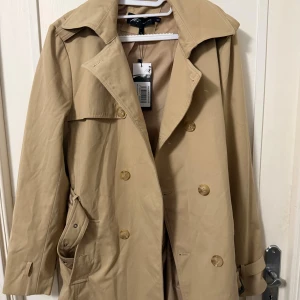 Beige trenchcoat från Mauvais, stl M - Stilren beige trenchcoat från Mauvais med klassisk dubbelknäppning, bred krage och snygga detaljer som bälte vid ärmslut och stora fickor. Perfekt för dig som vill ha en tidlös look med modern touch.