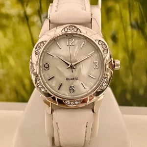 Vit klocka med glittriga detaljer - Snygg klocka med vit läderimitation och silverfärgat ur med glittrande stenar runt boetten. Urtavlan är pärlemorskimrande med tydliga siffror och markerade index. Perfekt för dig som gillar stilrena accessoarer med lite extra bling.Klockan är rustad med nytt batteri och fungerar fint!