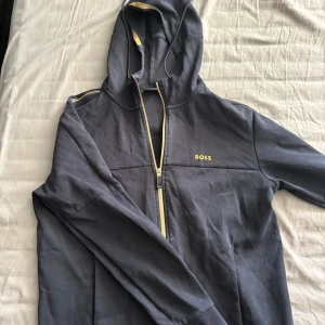 Mörkblå hoodie från BOSS med dragkedja - Snygg mörkblå hoodie från BOSS med gul logga på bröstet och gul detalj längs dragkedjan. Tröjan har huva med snörning och långa ärmar. Perfekt för chill eller när du vill ha en clean streetstil.