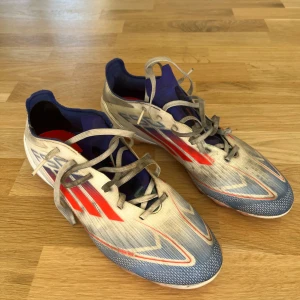 Adidas F50 fotbollsskor vit/blå/röd 44⅔ - Säljer ett par Adidas F50 fotbollsskor i storlek 44⅔. Skorna har en snygg design i vitt, blått och rött. Växtes snabbt ur. Konstgräsplanen har färgat dem lite svarta. Nypris 1500kr. En häl är förstärkt med hälskydd på insidan. 