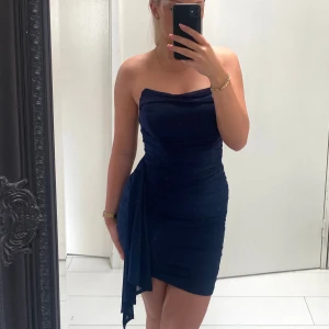 Marinblå axelbandslös klänning - Säljer en marinblå axelbandslös kortklänning i storlek 36 med tight passform och snygga draperingar. Klänningen har en asymmetrisk detalj i sidan. För referens är jag 164cm