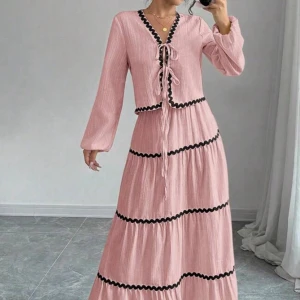 Lång rosa klänning med svarta detaljer - Supersöt långklänning i ljusrosa med svarta vågiga band längs volangerna. Klänningen har v-ringning med snörning framtill och långa ärmar. Perfekt för dig som gillar boho vibes och vill sticka ut med en unik look.