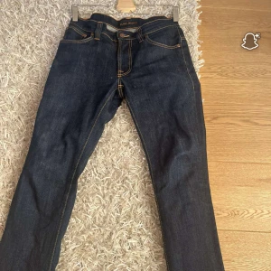 Mörkblå jeans från Nudie Jeans - Snygga mörkblå jeans från Nudie Jeans med klassisk femficksmodell och kontrastsömmar. Baksidan har de ikoniska vågformade broderierna på bakfickorna. Jeansen är raka i modellen och tillverkade i slitstarkt bomullstyg.