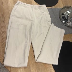 Beiga kostymbyxor från H&M, strl 34 - Snygga beiga kostymbyxor från H&M i storlek 34. Byxorna har raka ben, diskreta fickor fram och en enkel bakficka. Perfekta för en clean och stilren look. Materialet känns mjukt och lätt, vilket gör dem sköna att bära hela dagen.