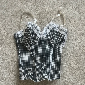 Svartvit rutig korsett med spets - Supersöt korsett i svartvitt rutigt mönster med vita spetsdetaljer och liten rosett framtill. Justerbara axelband och hakknäppning bak. Figurnära passform och lätt vadderade kupor för extra stöd. Perfekt för att lyfta din outfit med en retrovibe.