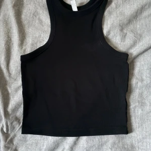Svart ribbad linnetopp från H&M Divided - Säljer en svart ribbad linnetopp från H&M Divided i storlek XS. Toppen är ärmlös med klassisk racerback och croppad passform. Perfekt att styla med jeans eller kjol för en enkel och clean look.