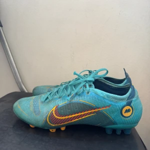 Fotbollskor Nike Mercurial 14 Pro AG - Säljer ett par turkosa Nike fotbollsskor med snörning och gula dobbar. Skorna har ett stort rött och gult Swoosh-märke på sidan och detaljer i gult. Ovandelen är i syntetmaterial och designen är sportig och modern, perfekt för dig som älskar fotboll. Skorna är i bra skick för att jag har varit skadad så jag har inte kunnat spela några matcher har bara tränat lite med dem därför säljer skorna pga att jag har slutat med fotboll. 
