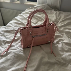 Ljusrosa handväska med silverdetaljer - Söt ljusrosa handväska i skinnimitation med silverfärgade nitar och dragkedjor. Väskan har både handtag och avtagbar axelrem, samt en liten ficka framtill med dragkedja. Perfekt storlek för det viktigaste och ger en cool touch till din outfit.