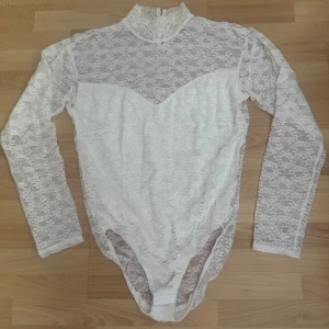 Vit spetsbody med lång ärm - Säljer en vit body med långärmade spetsdetaljer och högt hals. Hela toppen är täckt av blommig spets och har en snygg v-ringad illusion framtill. Bodyn stängs med tryckknappar nedtill och har en elegant, feminin vibe som passar perfekt till jeans eller kjol. Storlek lapp avklippt, M, hade på mig en gång, framsidan har foder, baksidan genomskinlig.
