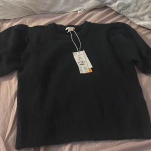 Mörkblå stickad topp med puffärm - Snygg mörkblå stickad topp med korta puffärmar och ribbade muddar. Modellen är croppad och har en enkel rund halsringning. Perfekt för dig som gillar en stilren men ändå trendig look. Tröjan har även  prislapp och är aldrig använd 