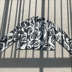 Svartvit mönstrad polotröja - Säljer en svartvit polotröja med coolt zebrainspirerat mönster. Tröjan är croppad och har långa ärmar samt en hög krage. Materialet är ribbat och känns mjukt mot huden. Perfekt för dig som vill sticka ut med en trendig look.