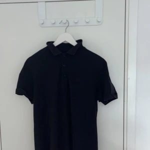 Svart kortärmad t-shirt från Hugo Boss - Svart t-shirt från Hugo Boss med korta ärmar och klassisk krage. Snygga Boss-loggor på bröstet och axeln, samt knappar framtill. Materialet är mjuk bomull som känns skönt mot huden. Perfekt för dig som gillar stilrena och enkla plagg med diskreta detaljer. Den är SLIM FIT 