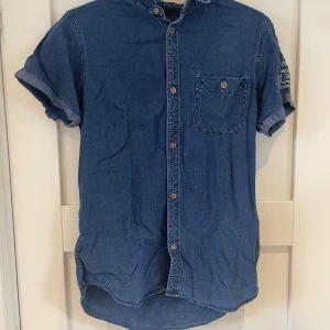 Blå jeansskjorta från Tifosi Label - Snygg blå jeansskjorta från Tifosi Label med korta ärmar och bröstficka. Skjortan har knappar framtill och broderad vit text på ena ärmen. Perfekt för dig som gillar avslappnad stil med denimkänsla.