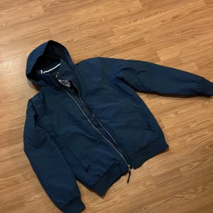 Blå vindjacka från Timberland med huva - Snygg blå vindjacka från Timberland med huva och dragkedja framtill. Jackan har ribbade muddar, två fickor och en diskret Timberland-logga på ärmen. Perfekt för blåsiga dagar och enkel att matcha med det mesta.