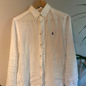 Vit linneskjorta från Ralph Lauren - Klassisk vit skjorta i linne från Polo Ralph Lauren med button down-krage och broderad marinblå logga på bröstet. Skjortan har långa ärmar och knäppning framtill med pärlemorskimrande knappar. Perfekt för en clean och stilren look.