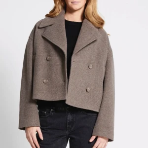 Beige croppad rock med knappar - Snygg croppad rock i beige med dubbelknäppning och bred krage. Jackan har en loose passform och långa ärmar, perfekt för dig som gillar stilren och trendig look. Tillverkad i ullblandning som håller dig varm under höst och vinter.