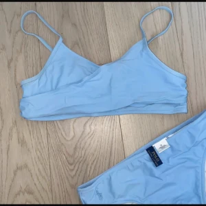 Ljusblå bikini från Polo Ralph Lauren - Superfin ljusblå bikini från Polo Ralph Lauren med enkel och stilren design. Toppen har smala axelband och omlott-detalj framtill. Bikinitrosan har broderad logga på sidan. Materialet är mjukt och stretchigt syntet, perfekt för sommardagar vid stranden.