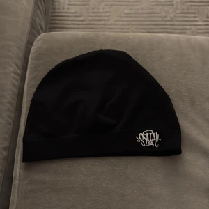 Syna World mössa  - Svart, slimmad mössa från Syna World med vit broderad logga framtill. Perfekt för kalla dagar och enkel att matcha med olika outfits. Tillverkad i mjukt syntetmaterial som sitter skönt på huvudet.