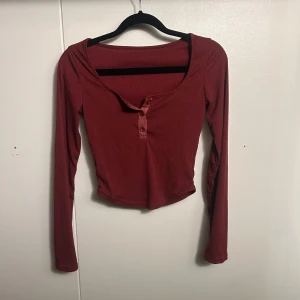 Ribbad vinröd långärmad topp - Säljer en vinröd ribbad topp med lång ärm och croppad passform. Toppen har en djup v-ringning med knappdetaljer framtill och är tillverkad i ett stretchigt material som sitter snyggt på kroppen. Perfekt för dig som gillar en enkel men trendig stil.