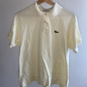 Vit pikétröja från Lacoste - Klassisk vit pikétröja från Lacoste med korta ärmar och den ikoniska krokodilloggan på bröstet. Tröjan har krage och knäppning med två knappar framtill. Tillverkad i mjuk bomull som känns skön mot huden. Perfekt för en clean och stilren look.