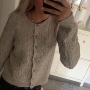 Beige kabelstickad kofta med knappar - Mysig beige kofta med kabelstickat mönster och rund hals. Koftan har knäppning framtill med små knappar och långa ärmar. Perfekt att slänga på sig när det är kyligt ute. Passar till allt och är superenkel att styla.