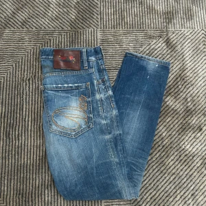 Dsquared2 blå jeans  - Snygga blå jeans från Dsquared2 med slitningar, stänkdetaljer och broderad logga på bakfickorna. Klassisk femficksmodell med raka ben och orange kontrastsömmar. Jeansen har en cool, använd look och är tillverkade i mjukt denimtyg.