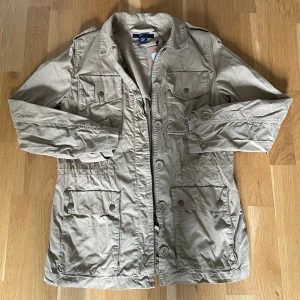 Beige jacka från Gant XS - Snygg beige parkajacka från Gant i bomull. Jackan har flera stora fickor med knappar, dragkedja och knäppning framtill samt klassisk krage. Perfekt för dig som gillar en stilren och avslappnad look.