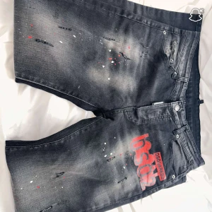 Svarta jeansshorts Dsquared2 - Snygga svarta jeansshorts från Dsquared2 med coolt rött DSQ2-tryck på framsidan. Shortsen har slitna detaljer och färgstänk i vitt och rött för en edgy vibe. Klassisk femficksmodell med knapp och dragkedja. Perfekt för dig som gillar streetstyle.