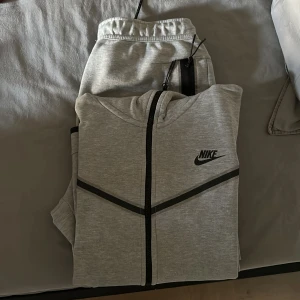 Nike tech fleece - Snygga ljusgrå mjukisbyxor från Nike med svarta dragkedjor på fickorna och elastisk midja med snörning. Perfekta för chill eller träning, med klassisk Nike-logga och mjukt material som känns skönt mot huden. Säljer koftan och byxorna för endast 700kr
