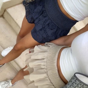 Beige Neo Noir kjol - Supersöt kort beige kjol med bred smock i midjan och dubbla volanger. Kjolen har en luftig och flowy känsla, perfekt för varma dagar. Den neutrala färgen gör den enkel att matcha med olika toppar och sneakers.