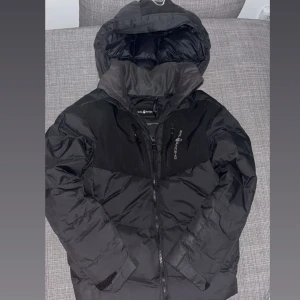 Svart pufferjacka från Sail Racing - Svart pufferjacka från Sail Racing med huva, dragkedja och flera fickor framtill. Jackan har en stilren look med logga på bröstet och patch på ärmen. Perfekt för kalla dagar och riktigt snygg. Hör av er vid bud!