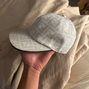 Vit Fendi keps med logomönster - Snygg vit keps från Fendi med deras ikoniska FF-logomönster över hela kepsen. Kepsen har böjd skärm med svart kant och är tillverkad i 100% polyester. Perfekt för dig som vill ha en clean och lyxig look med designer vibe.