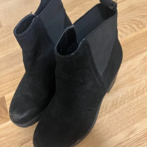 Svarta mocka chelsea boots med klack - Snygga svarta chelsea boots i mocka med bred klack och rund tå. Bootsens elastiska paneler på sidorna gör dem enkla att dra på och av. Perfekta för dig som gillar stilrena och klassiska skor med lite höjd. Passar till både jeans och kjol.