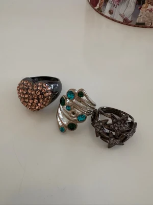 Tre statementringar med stenar och stjärnor - Säljer tre chunky ringar: en svart ring med hjärta täckt av glittrande bronsfärgade stenar, en silvrig ring med blå och gröna stenar i vågig form samt en mörk ring med flera stjärnor dekorerade med små kristaller. Perfekta accessoarer för att sticka ut.