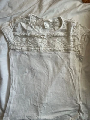 Vit spetsblus från H&M XS - Säljer en vit blus från H&M i storlek XS. Toppen har korta ärmar och vacker spets upptill med broderade detaljer. Rund halsringning och mjukt bomullstyg nedtill. Perfekt för dig som gillar romantisk stil och ljusa färger. En liten fläk på tröjan som knappt syns men inte går bort..