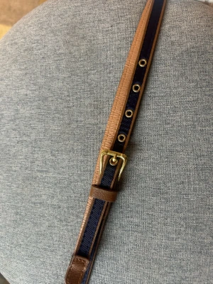 Brunt och marinblått bälte - Snyggt bälte i brunt skinn med marinblå detalj och guldfärgat spänne. Används inte! ⚠️Största omkretsen är ca 79 cm och minsta 69 cm